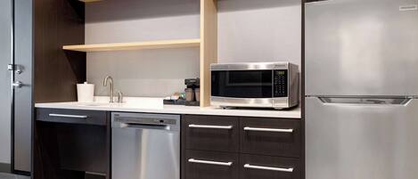 A modern kitchen with a built-in dishwasher, a stainless steel refrigerator, a microwave, and wooden shelves above the countertop.