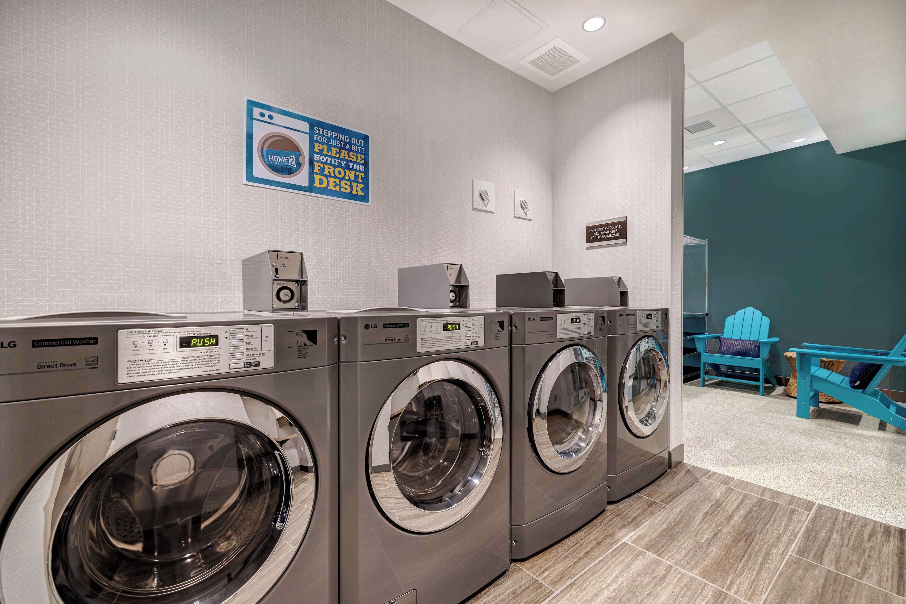 A laundry facility with three commercial washers and dryers, a sign instructing to notify the front desk before stepping out, and a blue chair in the background.