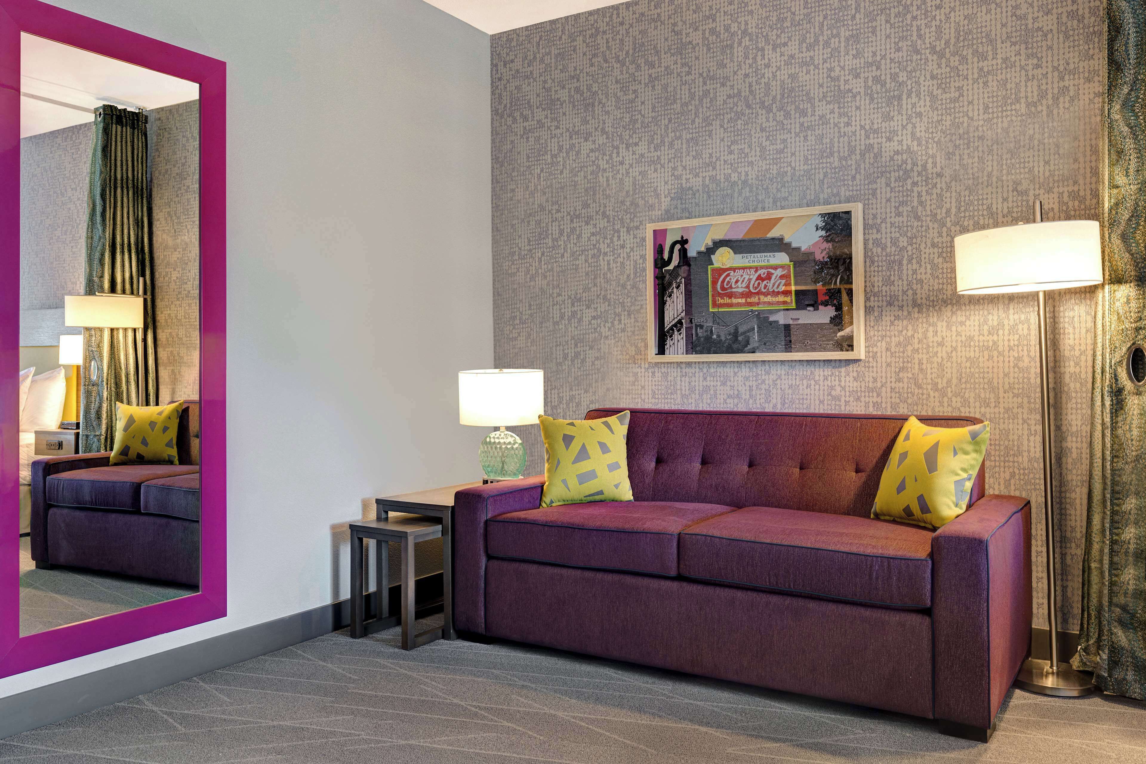 A modern hotel room with a purple sofa, two lamps, a framed picture on the wall, and a large mirror.