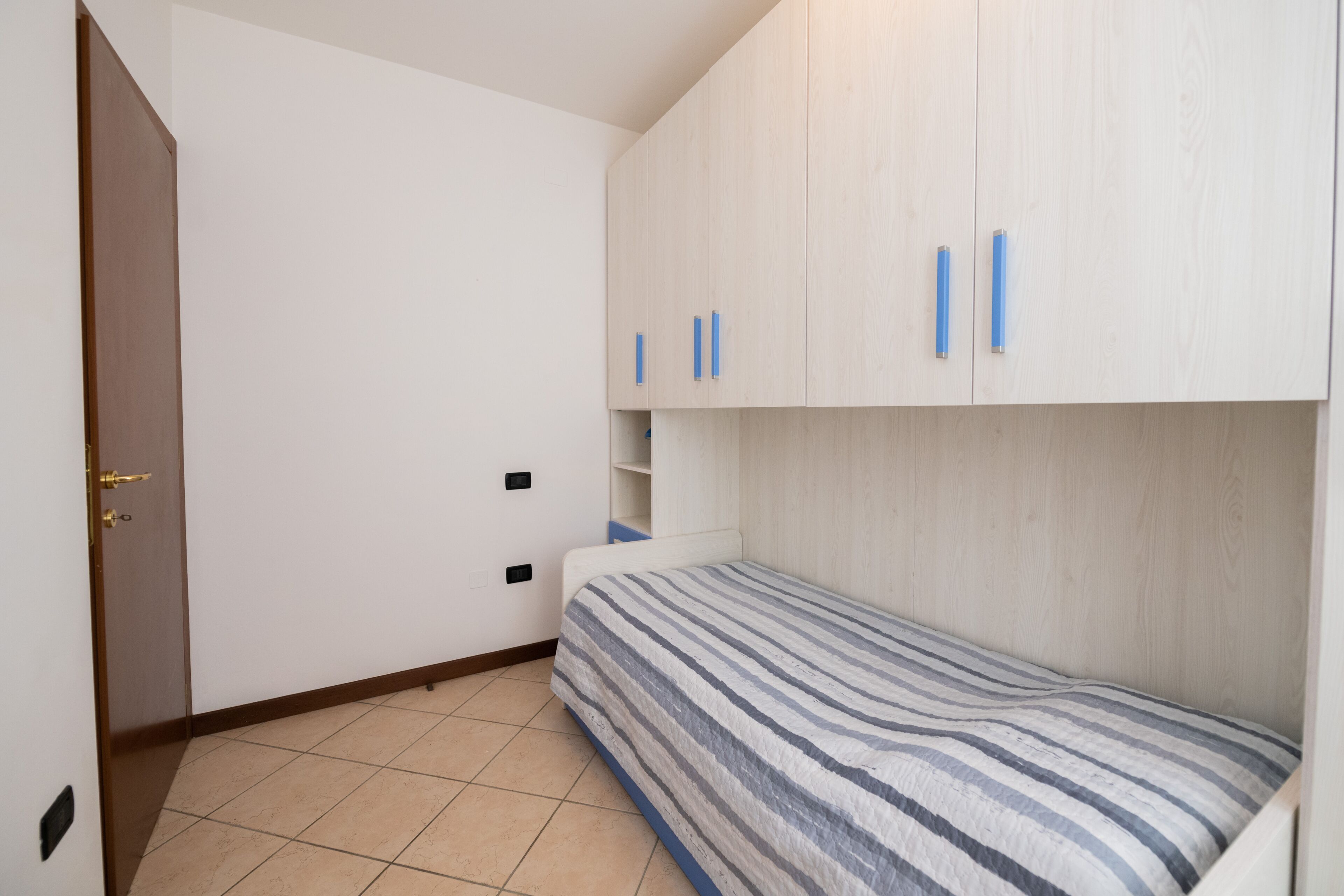 Ein kleines Zimmer mit einem Einzelbett, einem Holzschrank und einer Tür mit Messingbeschlag.