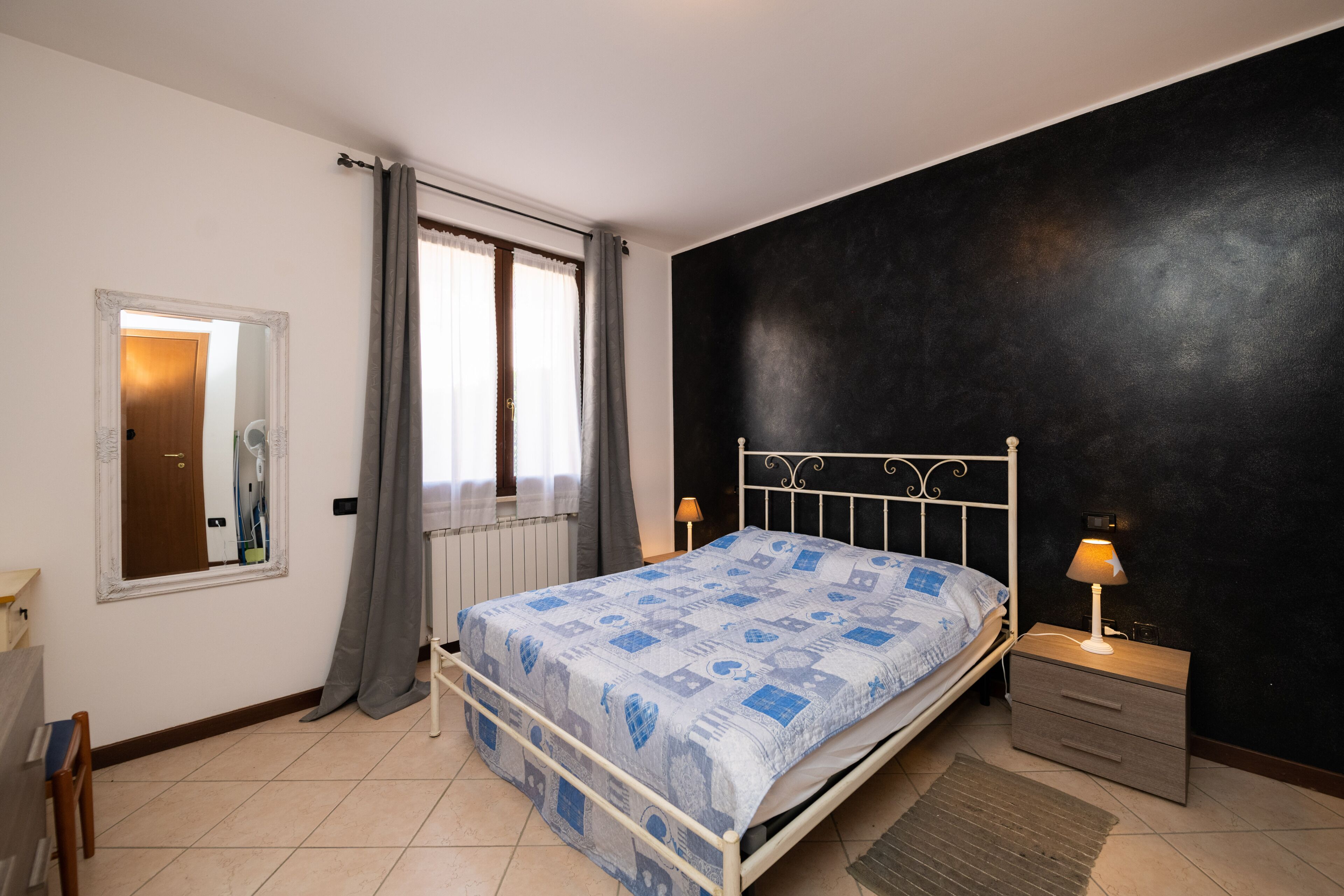 A bedroom with a bed, a nightstand, a mirror, a window with curtains, and a radiator.