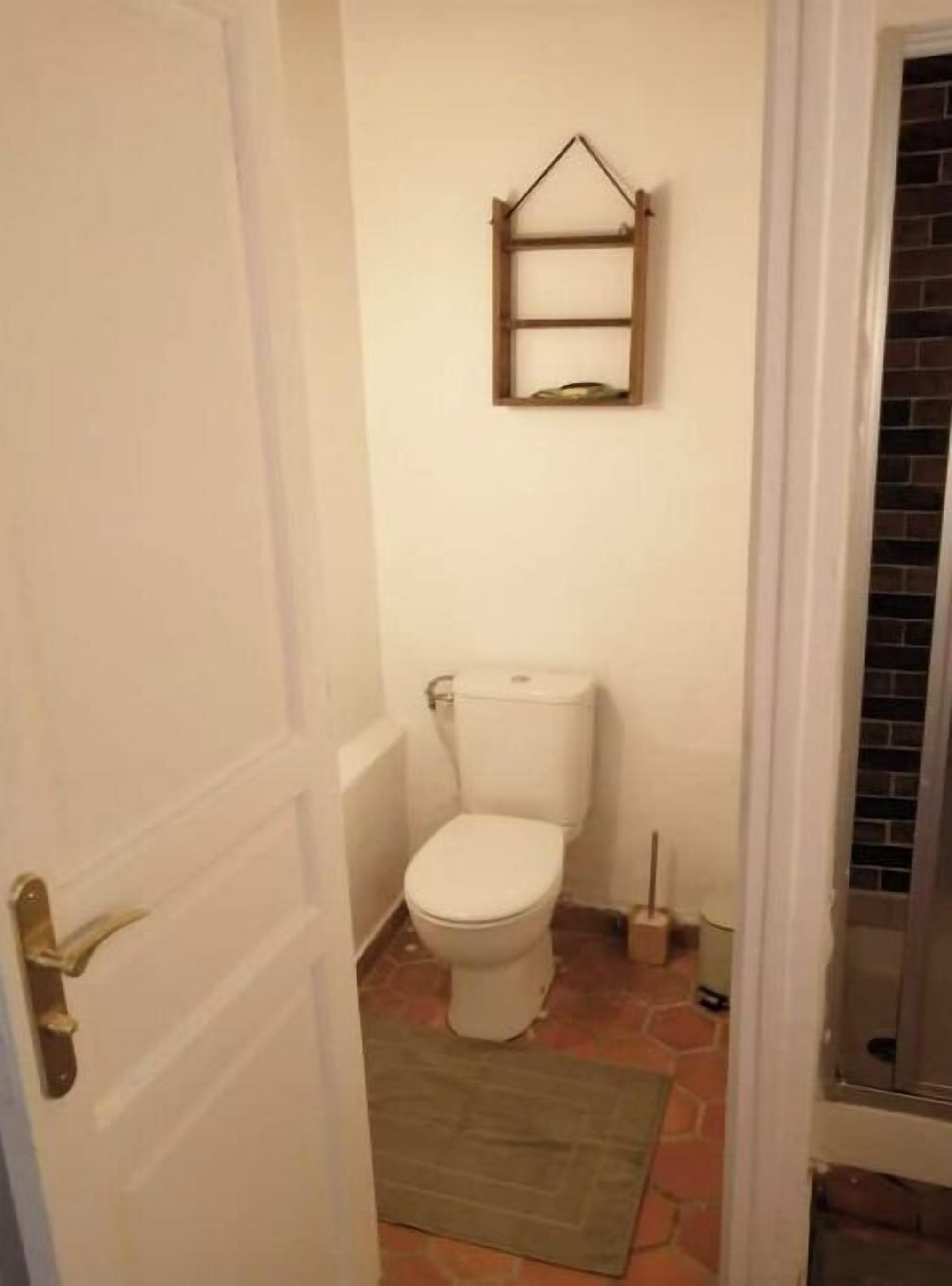 A small bathroom with a toilet, a wooden shelf above it, and a door with a brass handle.
