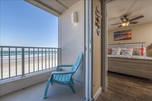 A balcony with a view of the beach, a blue Adirondack chair, and a bedroom with a bed and a ceiling fan.