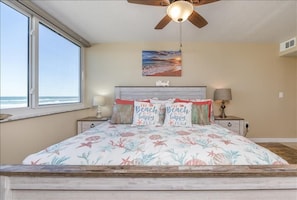 A bedroom with a large bed, a ceiling fan, a window with a view of the ocean, and a painting on the wall. - Welcome to Beach Life! 106 South Point " traffic free beach" (Ponce Inlet)