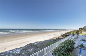 A beach with a clear sky, a sandy shore, and a few people walking. There is a white railing and some greenery in the foreground. - Welcome to Beach Life! 106 South Point " traffic free beach" (Ponce Inlet)
