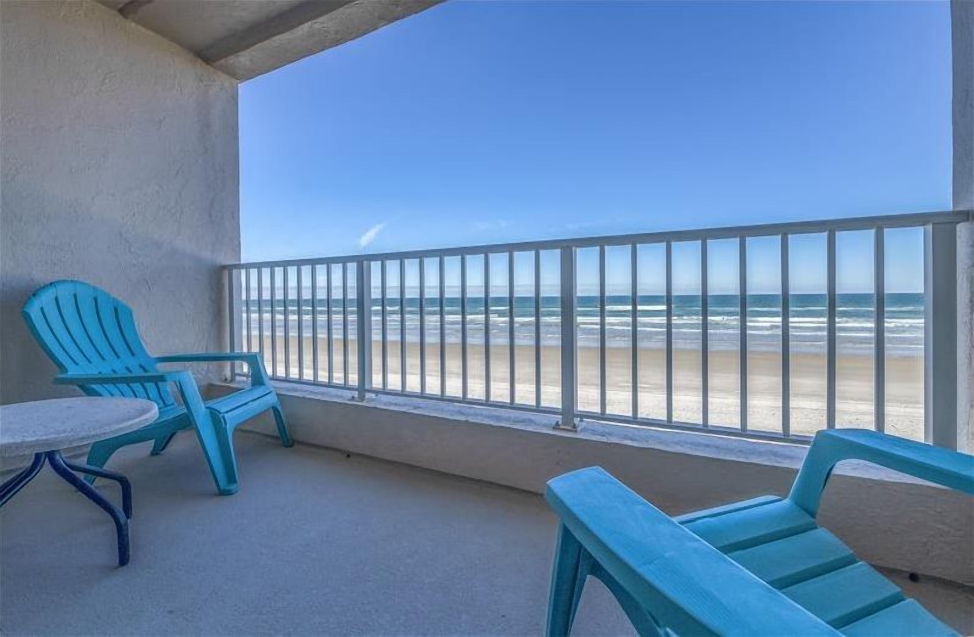 Een balkon met uitzicht op het strand, met twee blauwe stoelen en een kleine ronde tafel.