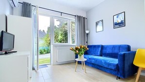 A modern living room with a blue sofa, a yellow chair, a TV on a white cabinet, and a window with a view of greenery. - Appartment B1 Sarbinowo, 200 m vom Strand und Centrum (Sarbinowo)