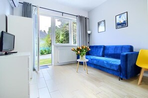 A modern living room with a blue sofa, a yellow chair, a TV on a white cabinet, and a window with a view of greenery.