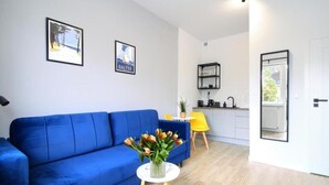 Una sala de estar moderna con un sofá azul, una mesita blanca con un jarrón de flores y una zona de cocina al fondo.