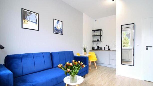 A modern living room with a blue sofa, a small white table with a vase of flowers, and a kitchen area in the background. - Appartment B1 Sarbinowo, 200 m vom Strand und Centrum (Sarbinowo)