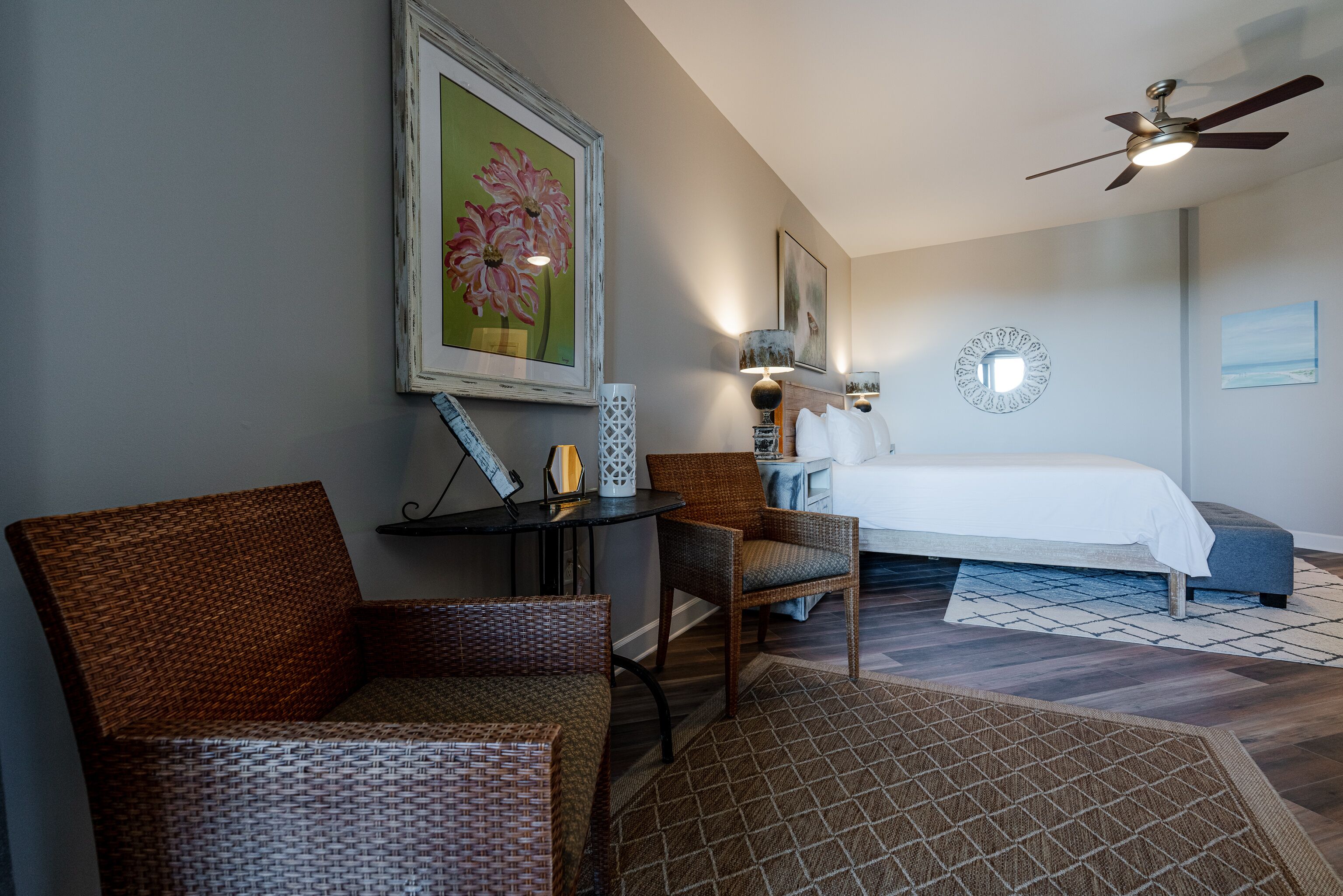 A bedroom with a bed, two wicker chairs, a small table, and a ceiling fan.