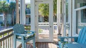 Eine überdachte Veranda mit zwei blauen Adirondack-Stühlen, einer weißen Tür und Blick auf Strand und Palmen.