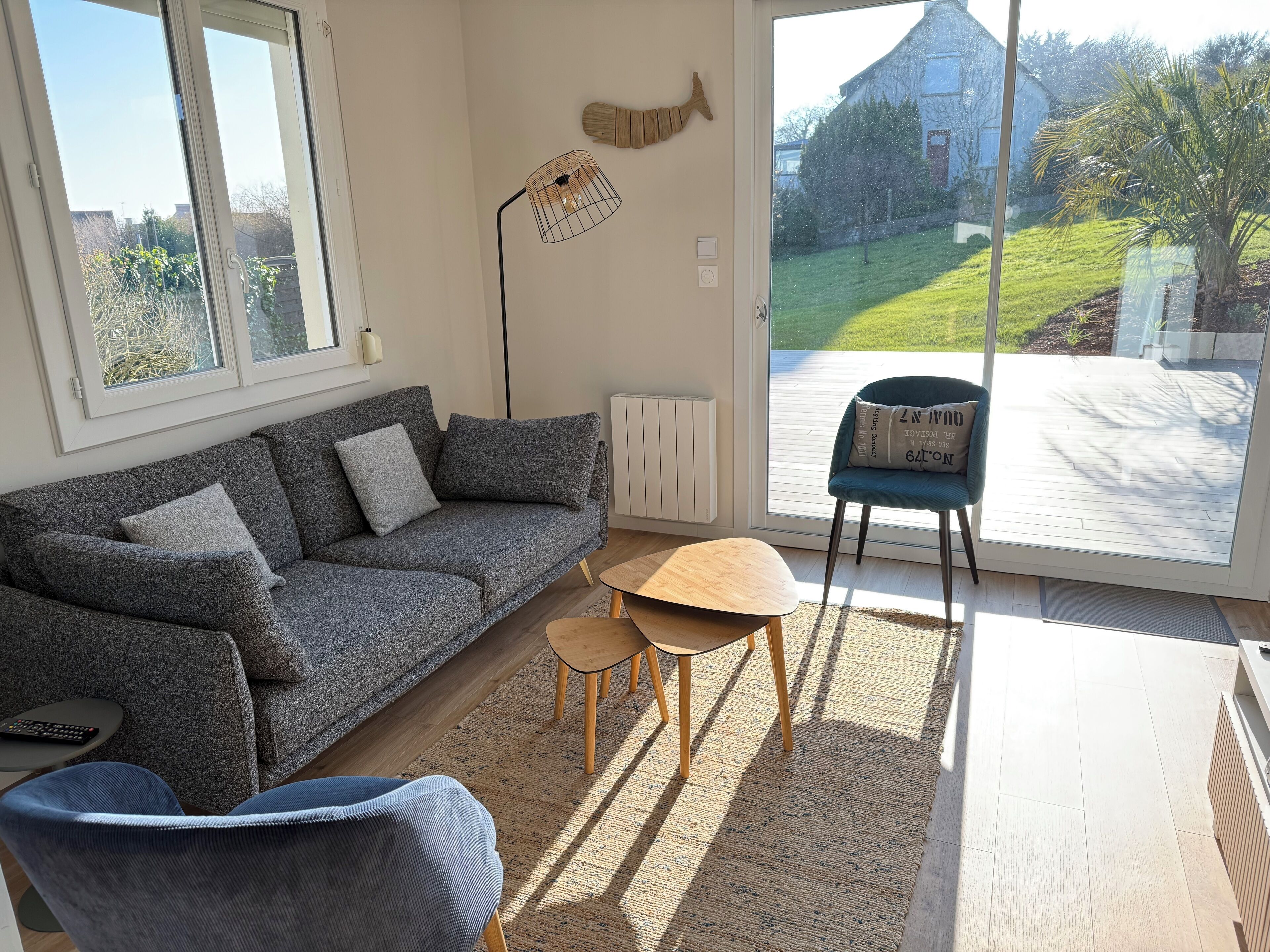A modern living room with a grey sofa, a blue armchair, a small wooden table, and a view of a garden through large glass doors.