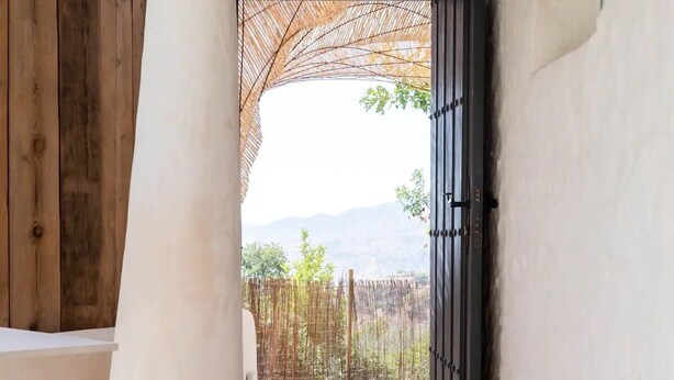 A room with a wooden bench, a concrete pillar, a black door, and a view of a garden with a fence and trees. - Kaolin Rocabella Apartment with Private Sandy Pool (El Chorro)