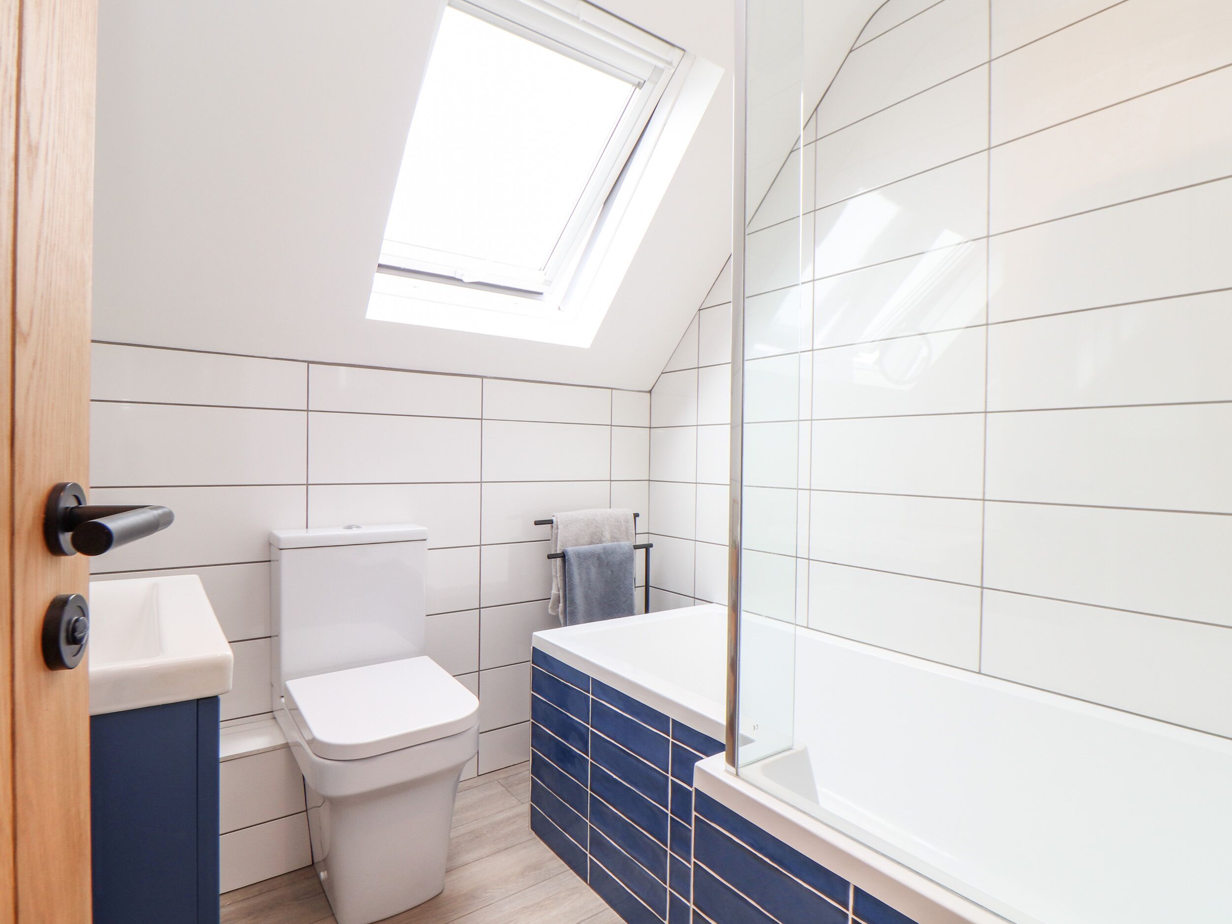 A modern bathroom with a skylight, white tiles, a toilet, and a blue-tiled shower area.