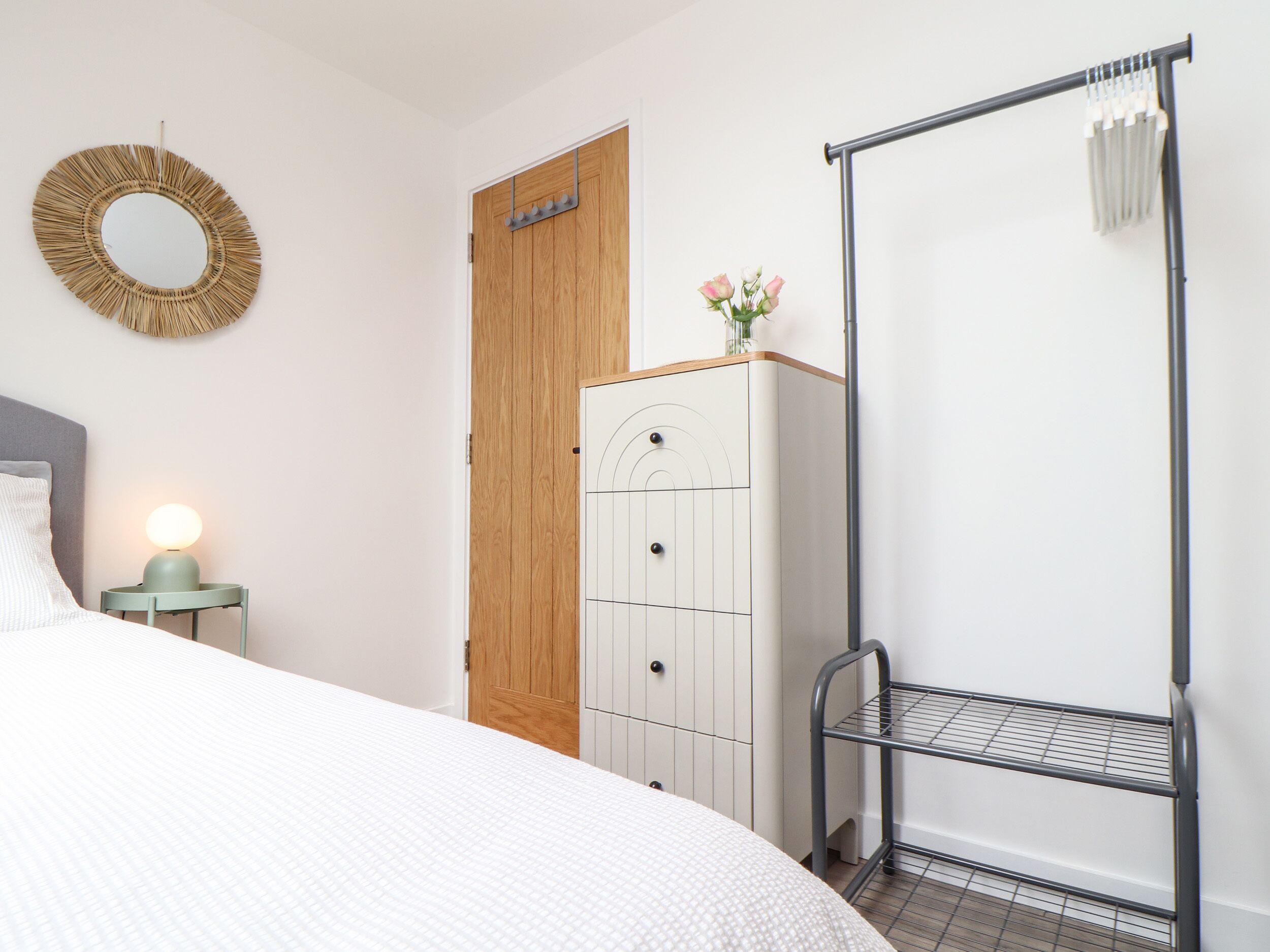 A bedroom with a bed, a dresser, a wardrobe, a hanging rack, and a wall-mounted mirror.