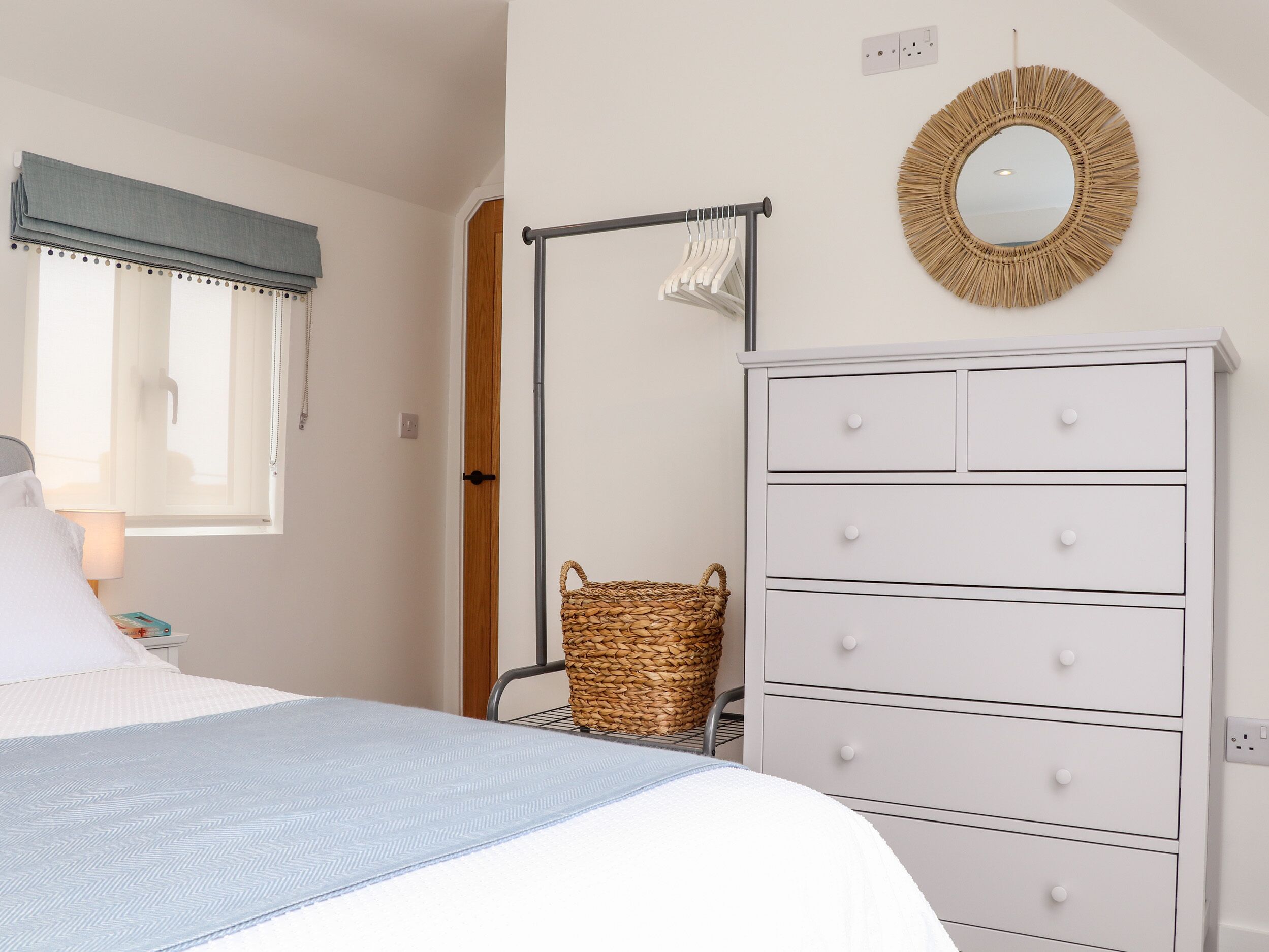 Quarto com cama, cómoda com gavetas, cabideiro, cesto de vime e espelho na parede.