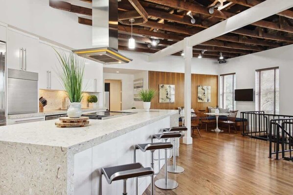 A modern kitchen with a central island, bar stools, and a dining area with a high ceiling and exposed beams.