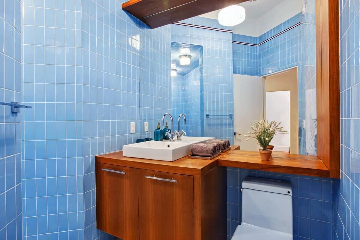 A bathroom with blue tiles, a wooden vanity, a white sink, two faucets, a mirror, and a potted plant.