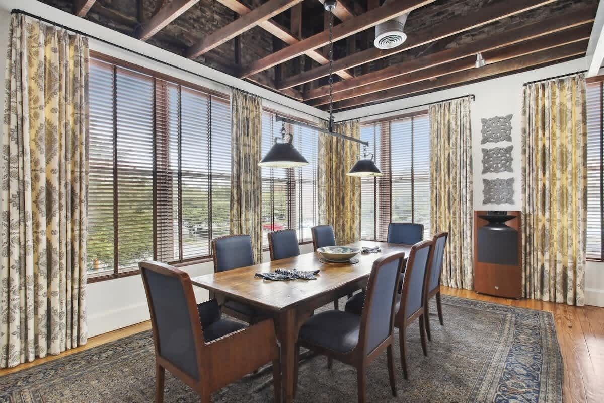 Un comedor con una mesa de madera, seis sillas, dos lámparas colgantes y ventanas grandes con cortinas.