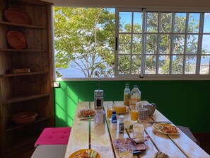 Taula de dinar amb menjar per esmorzar, finestra amb vista a arbres i prestatgeria de fusta amb plats.