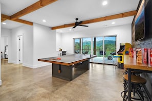 Relax in a serene space with a ping pong table, high ceiling, and large windows offering a view of lush greenery.