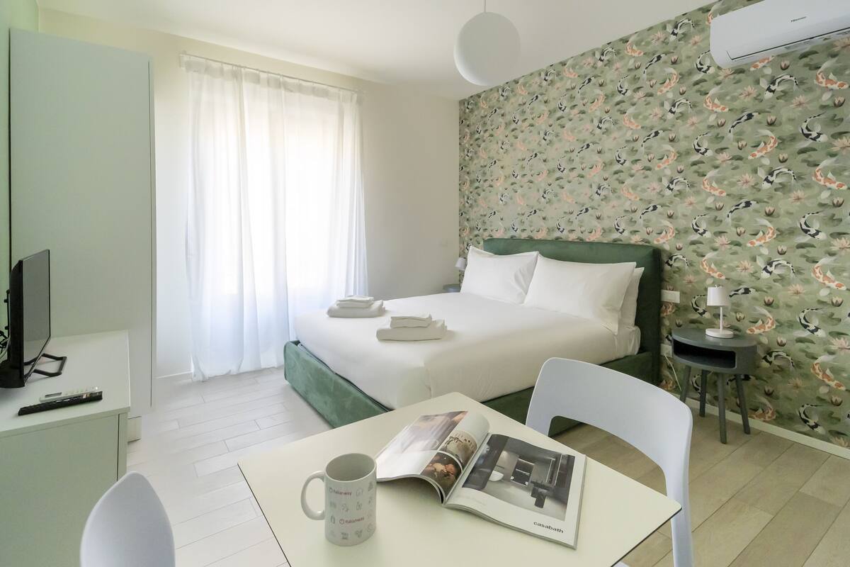 A hotel room with a bed, a desk with a chair, a TV, and a magazine on the table.