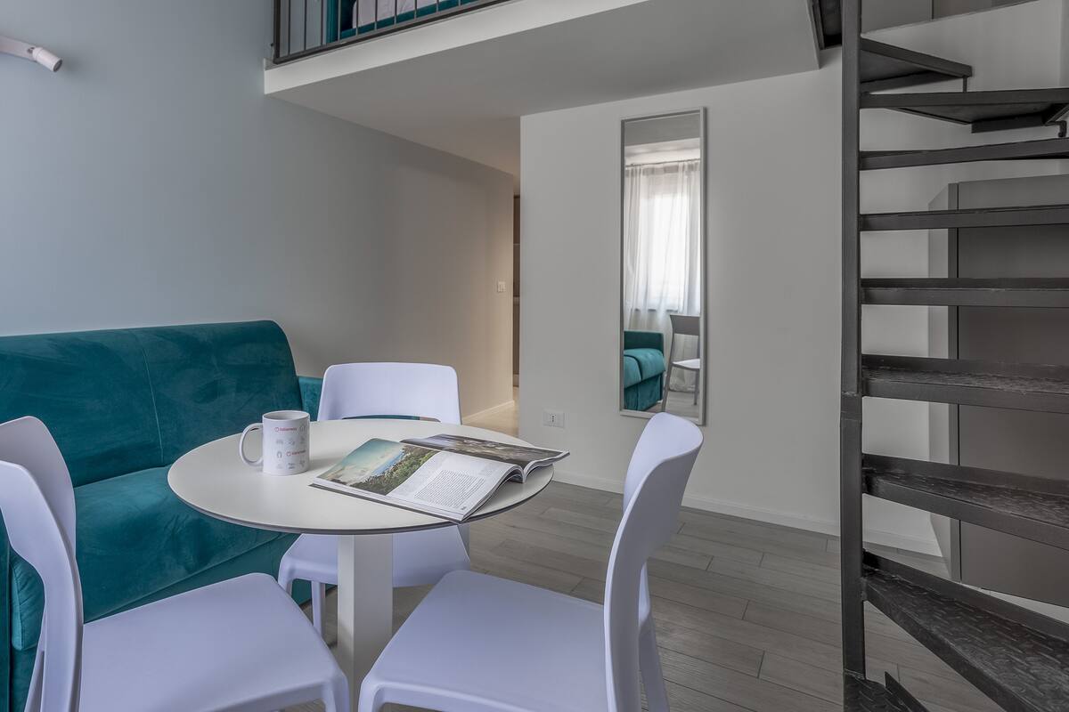A modern room with teal upholstered seating, a small round table with a magazine, and a white chair. A mirror reflects a teal sofa and a window with curtains.