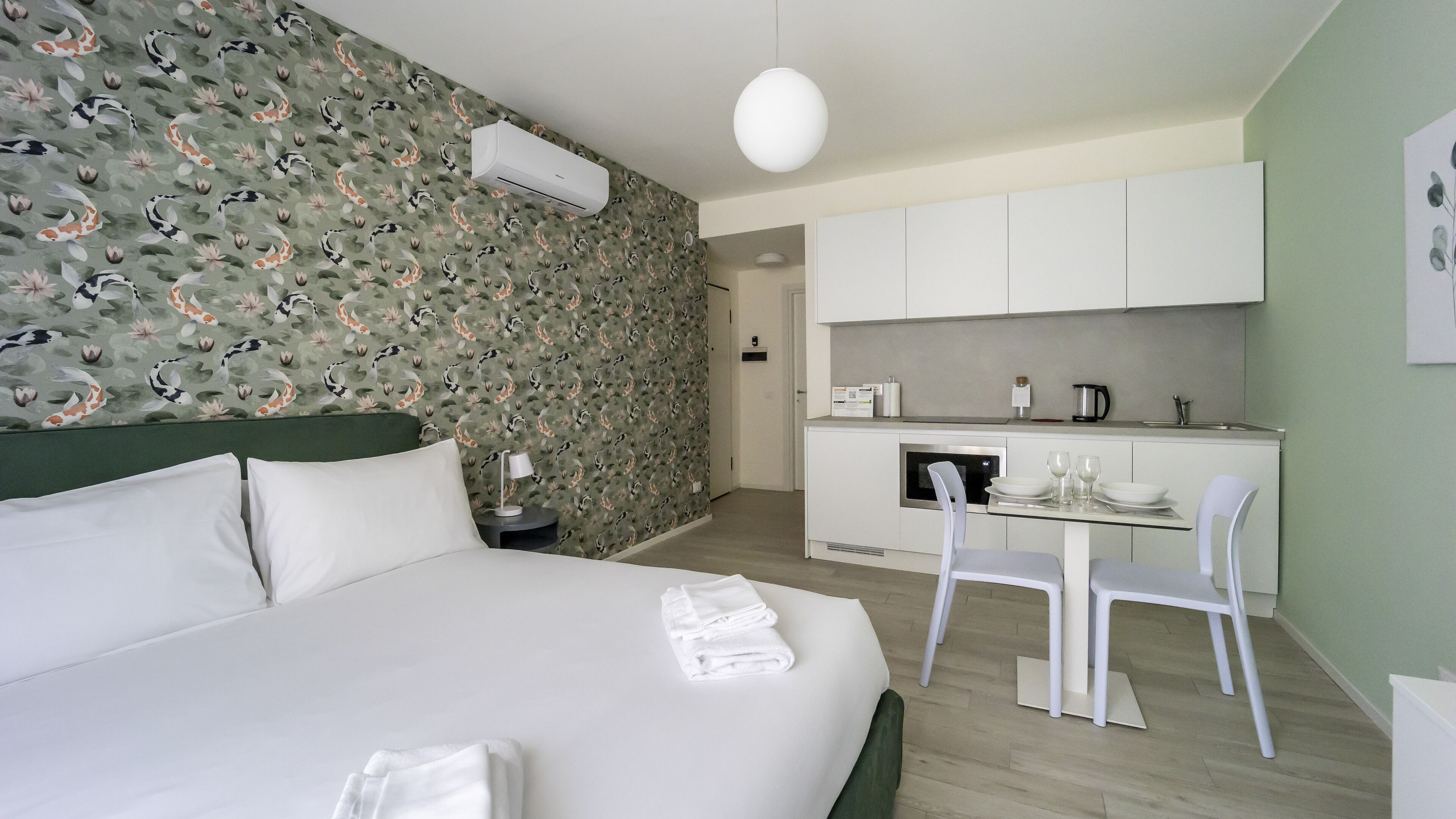 Hotellihuone, jossa on sänky, pieni pöytä tuoleineen, pieni keittiö ja kylpyhuone.