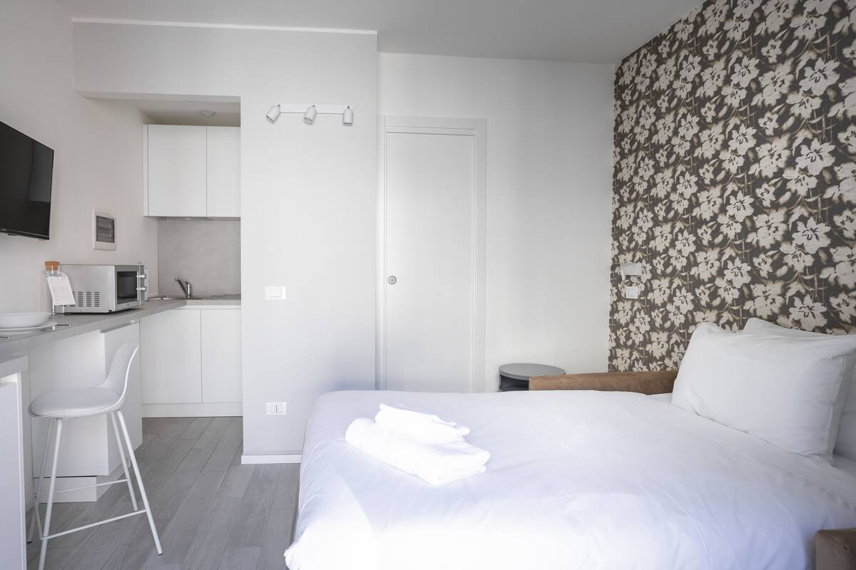 A modern hotel room with a kitchenette, a bed with white linens, and a floral-patterned wall.