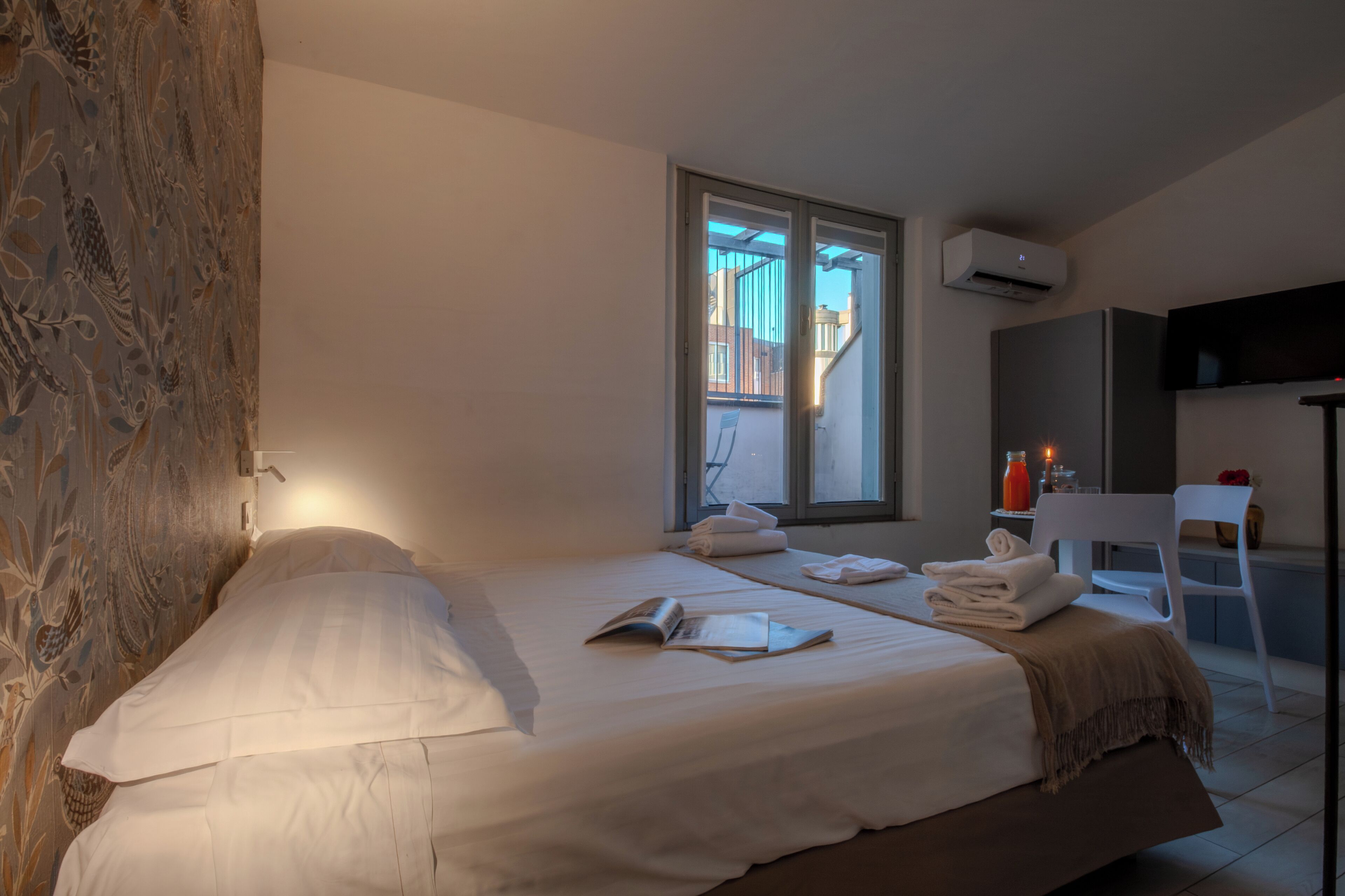 A hotel room with two beds, a window with a view of the outside, a flat-screen TV mounted on the wall, and a small table with a chair.
