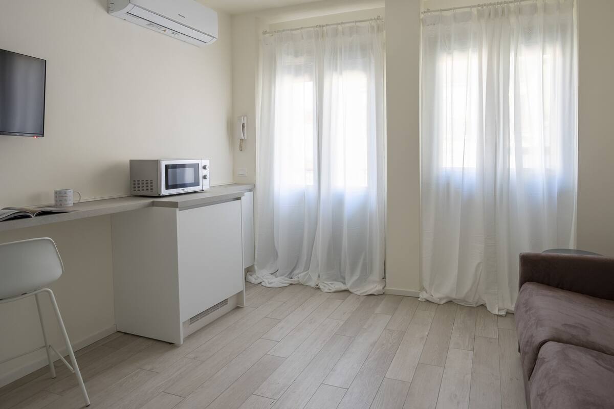 A modern, minimalistic living space with a kitchenette, a flat-screen TV, a sofa, and sheer curtains.