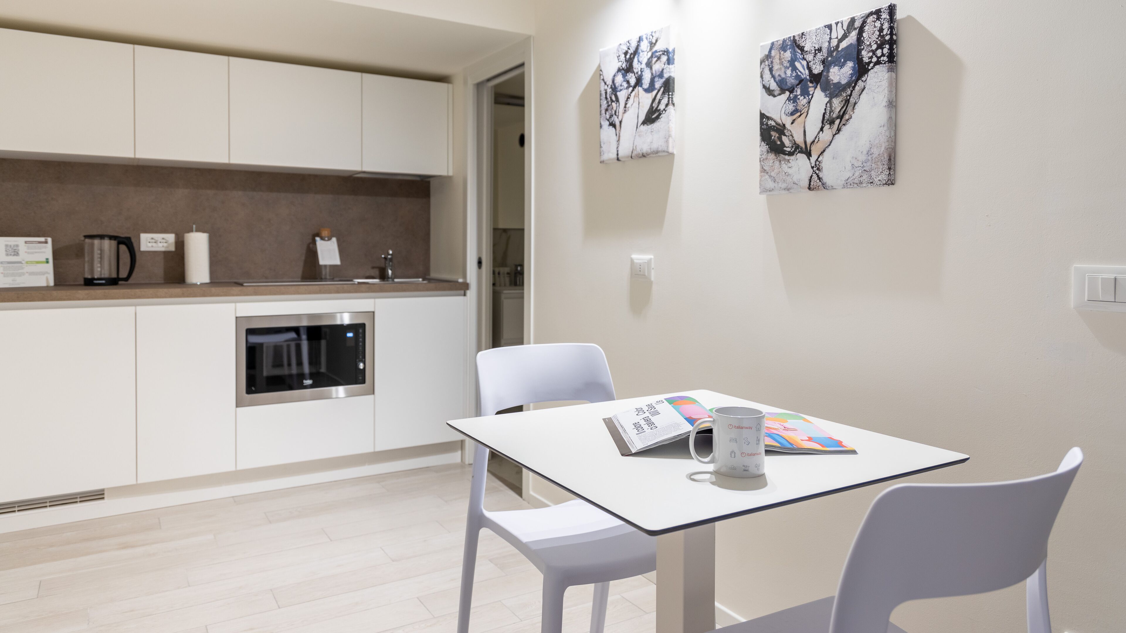 Una cocina moderna con armarios blancos, horno integrado y cafetera. Hay una mesa con una revista y una taza encima.