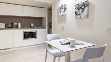 Una cocina moderna con armarios blancos, horno integrado y cafetera. Hay una mesa con una revista y una taza encima.