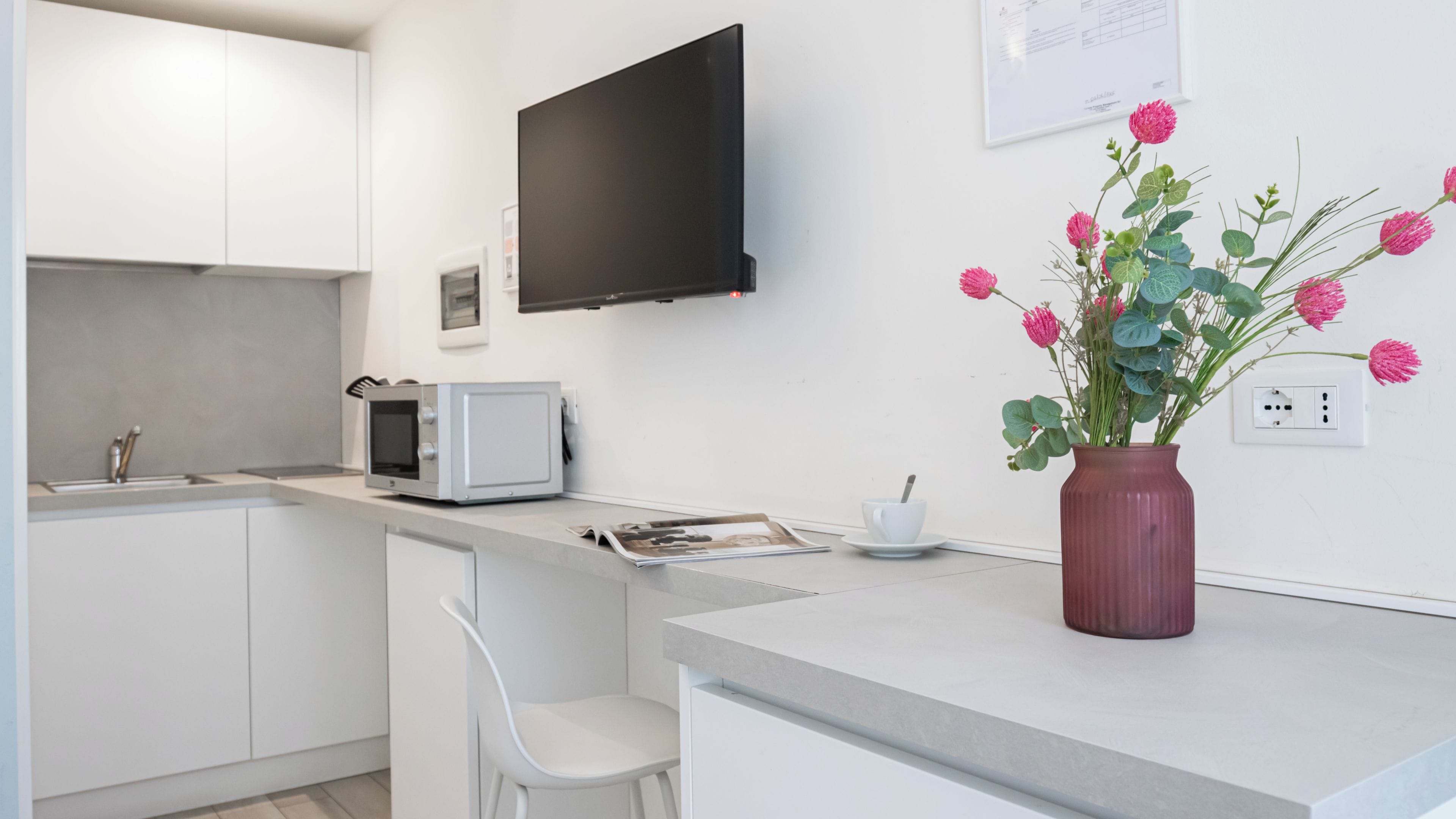 A modern kitchen with white cabinets, a microwave, a flat-screen TV, and a vase with flowers on the counter.