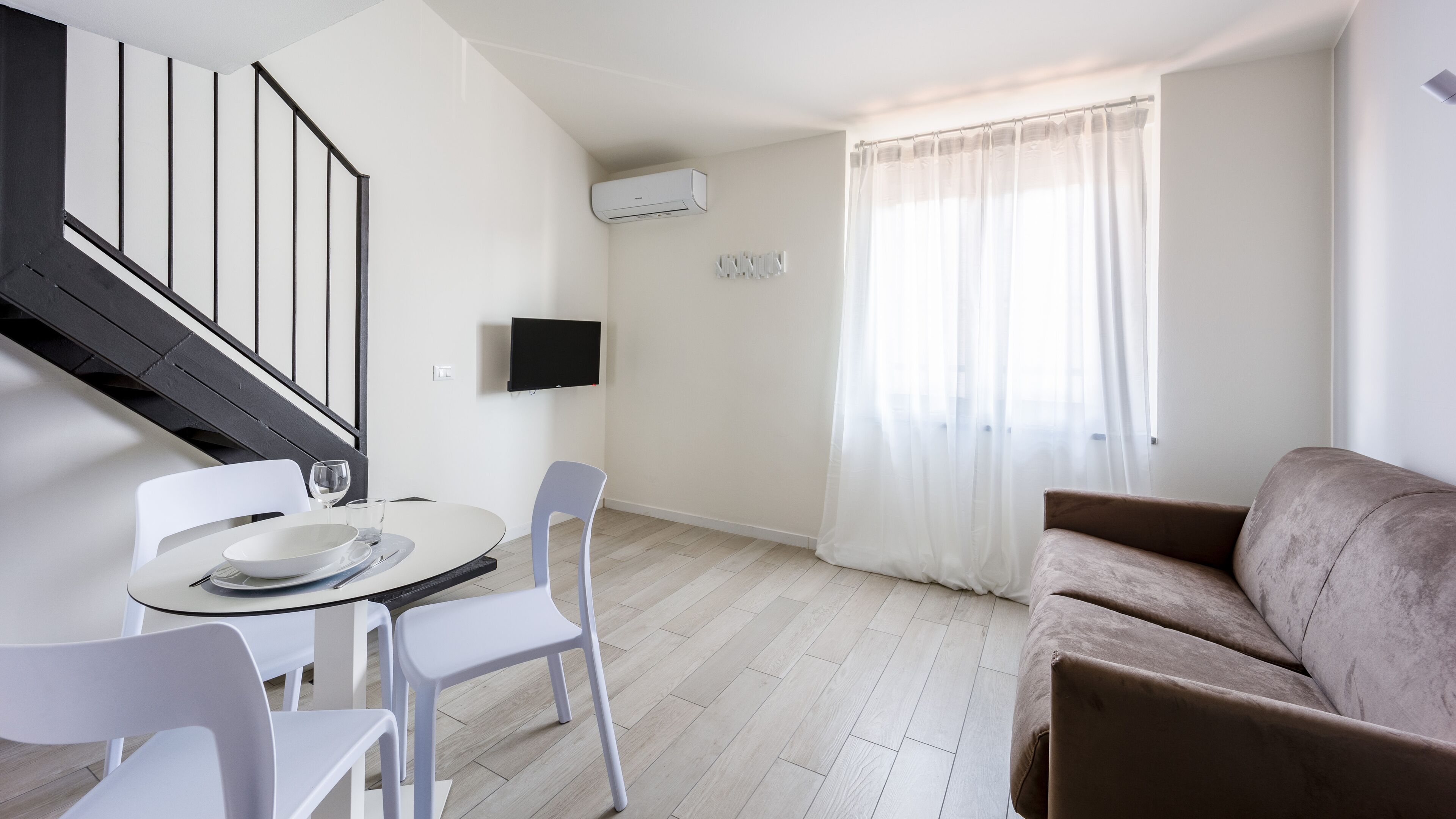 Een moderne woonkamer met een trap, een eethoek met een tafel en stoelen, een flatscreen televisie aan de wand en een groot raam met dunne gordijnen.