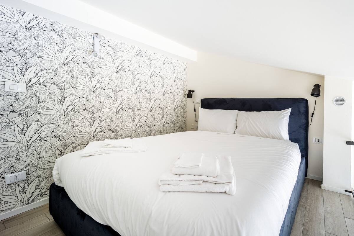 A neatly made bed with white linens and folded towels, a dark headboard, and a wall with a botanical wallpaper pattern.