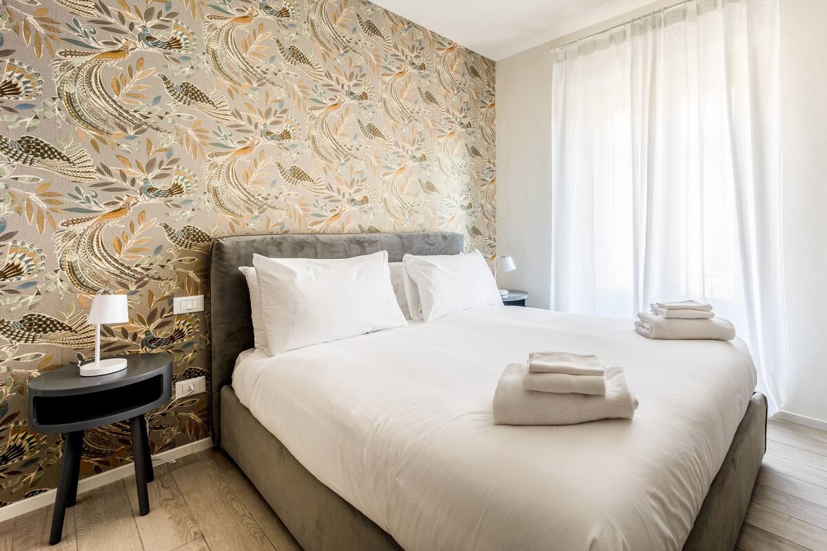 A hotel room with a large bed, a bedside table with a lamp, and a wall with a floral pattern.