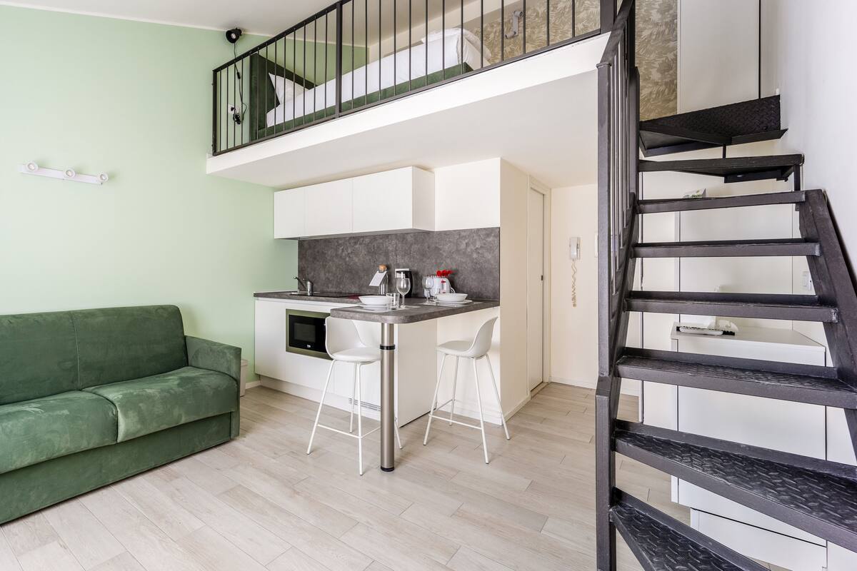 A compact living space with a green sofa, a kitchenette, a dining area, and a staircase.