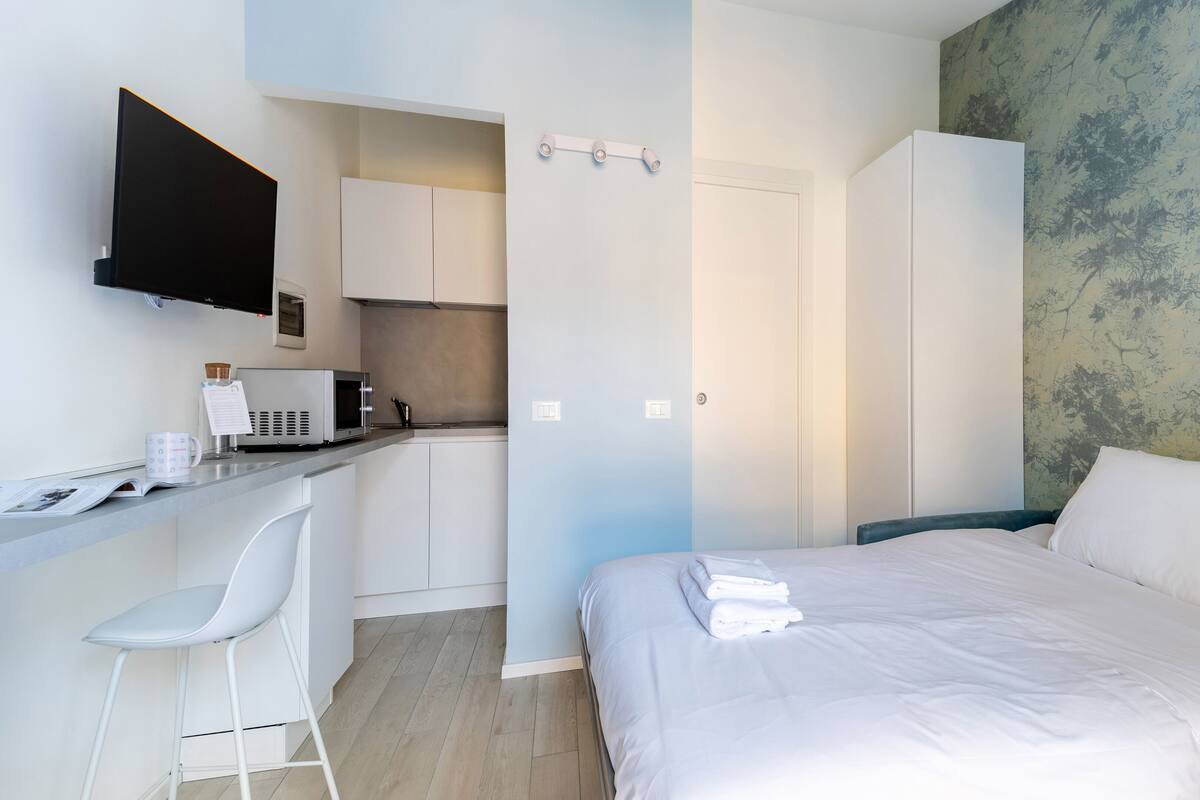 A modern bedroom with a bed, a desk, and a kitchenette.