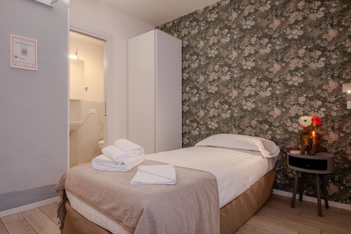 A hotel room with a bed, a nightstand, a floral wallpaper, and a door leading to a bathroom.