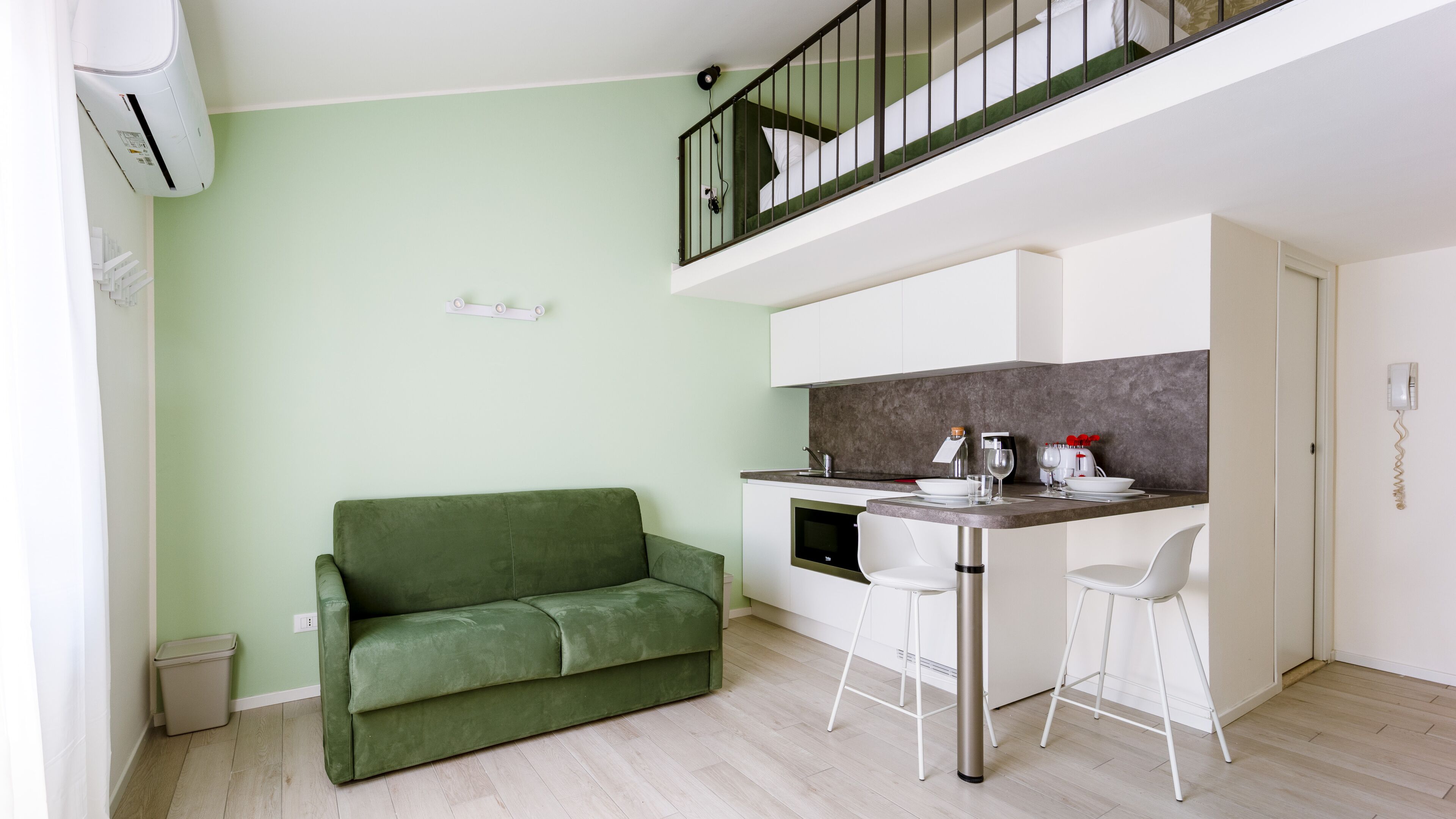 Un espacio habitable compacto con kitchenette, un sofá verde y una pequeña zona de comedor con dos taburetes tipo bar.