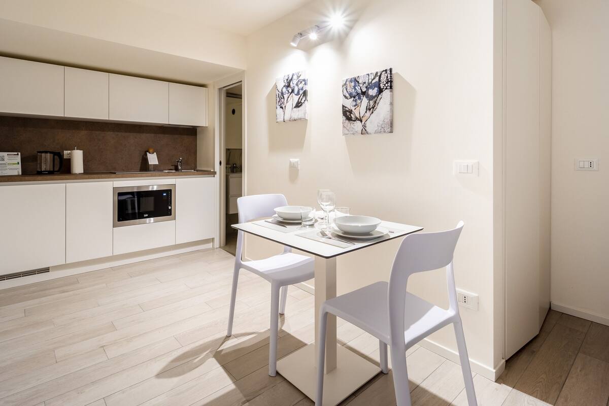 A modern kitchen with white cabinetry, a built-in microwave, and a dining area with a glass table and white chairs.