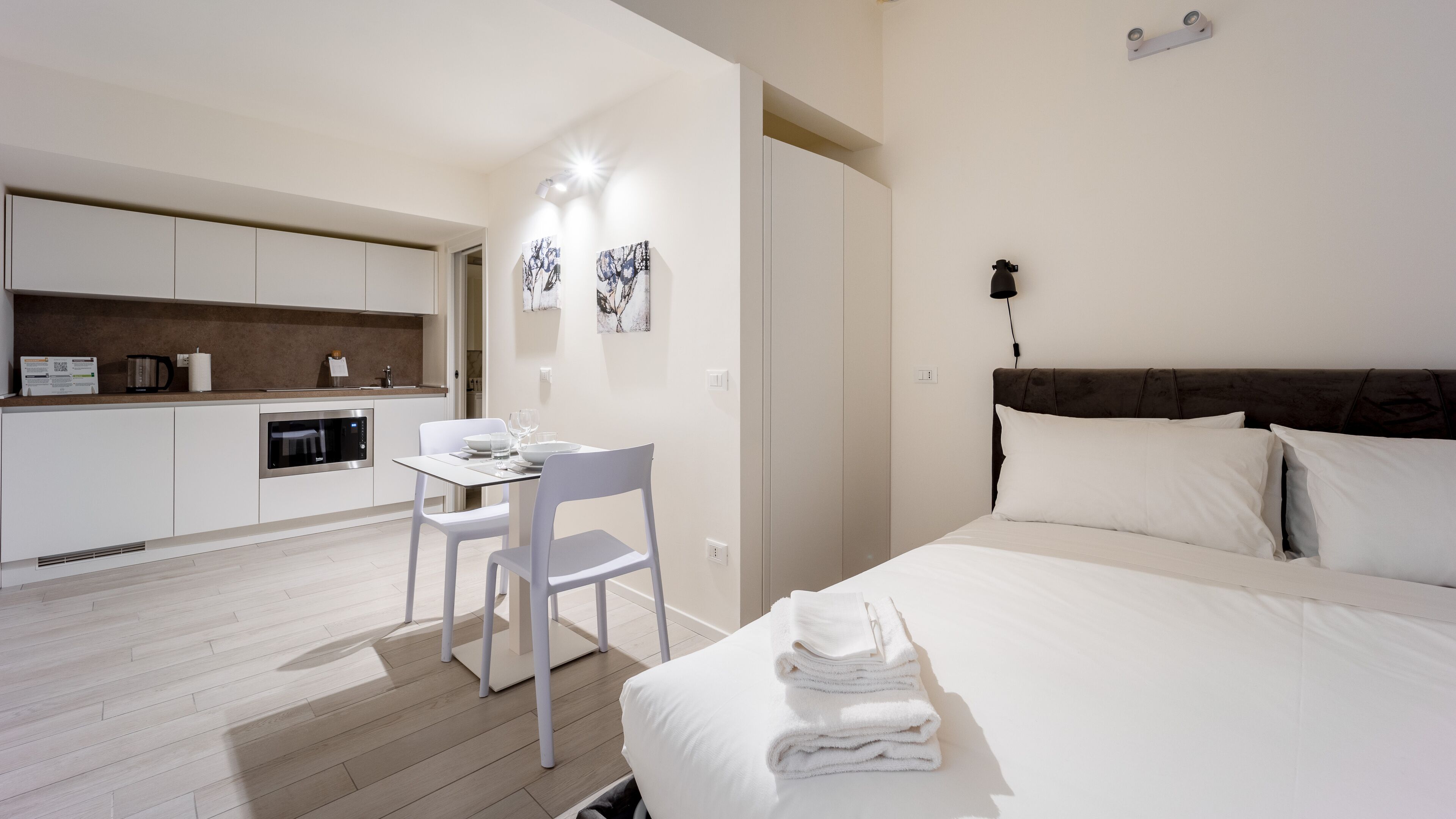 A modern bedroom with a large bed, a desk with a chair, a kitchen area with cabinets and appliances, and artwork on the walls.