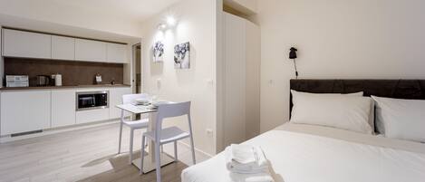 A modern bedroom with a large bed, a desk with a chair, a kitchen area with cabinets and appliances, and artwork on the walls.