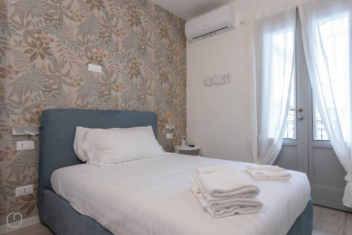 A neatly made bed with white linens, a grey headboard, and a floral-patterned wallpaper. There is a white bedside table with a phone and a small lamp. The room has a door with a white curtain and a wall-mounted air conditioning unit.