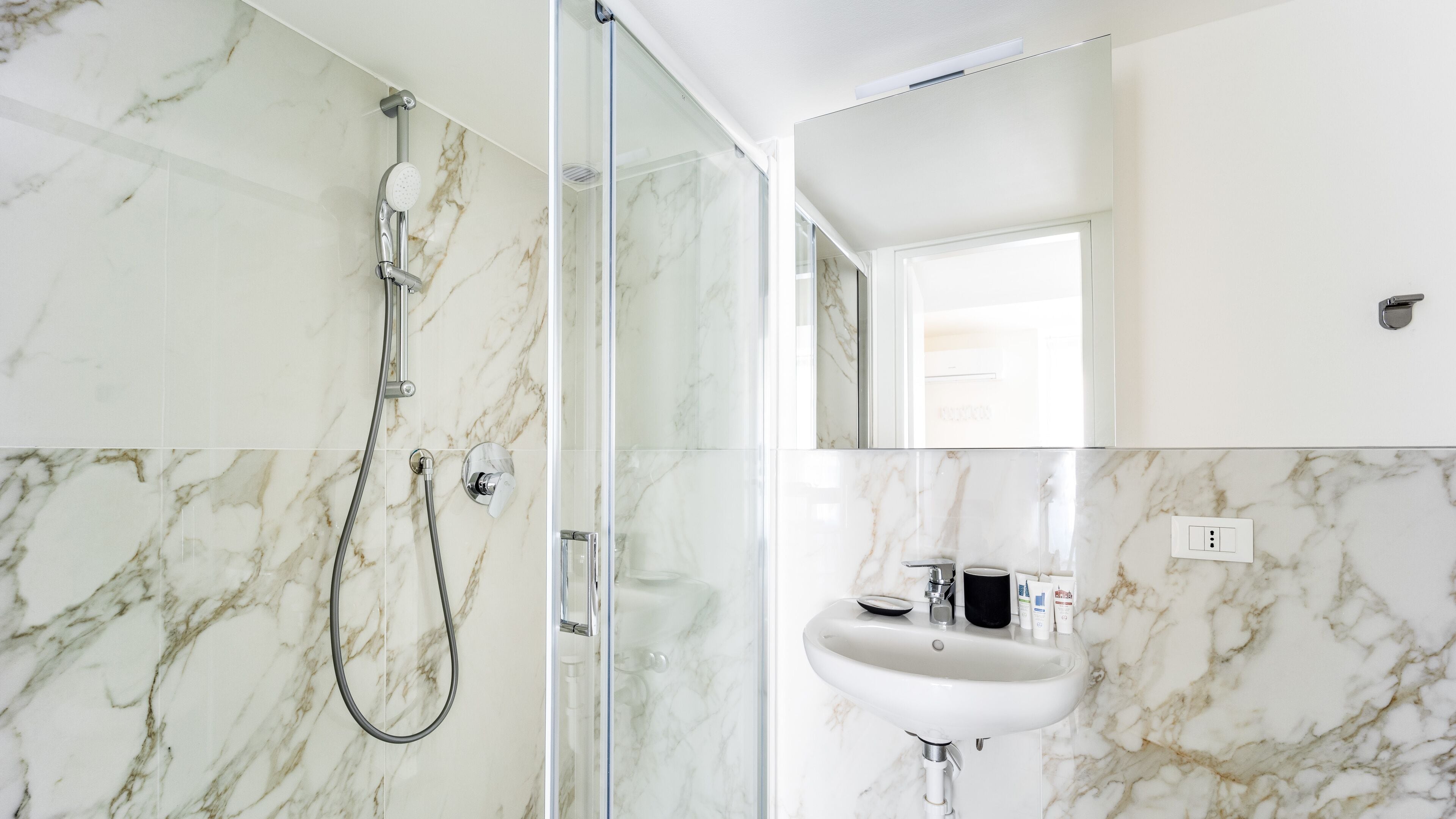 Un baño moderno con ducha envuelta en vidrio, lavamanos blanco y paredes de mármol.