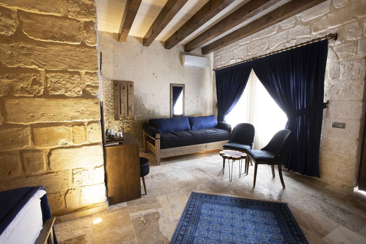 A rustic room with exposed stone walls, a wooden ceiling, a sofa, two chairs, and a patterned rug.