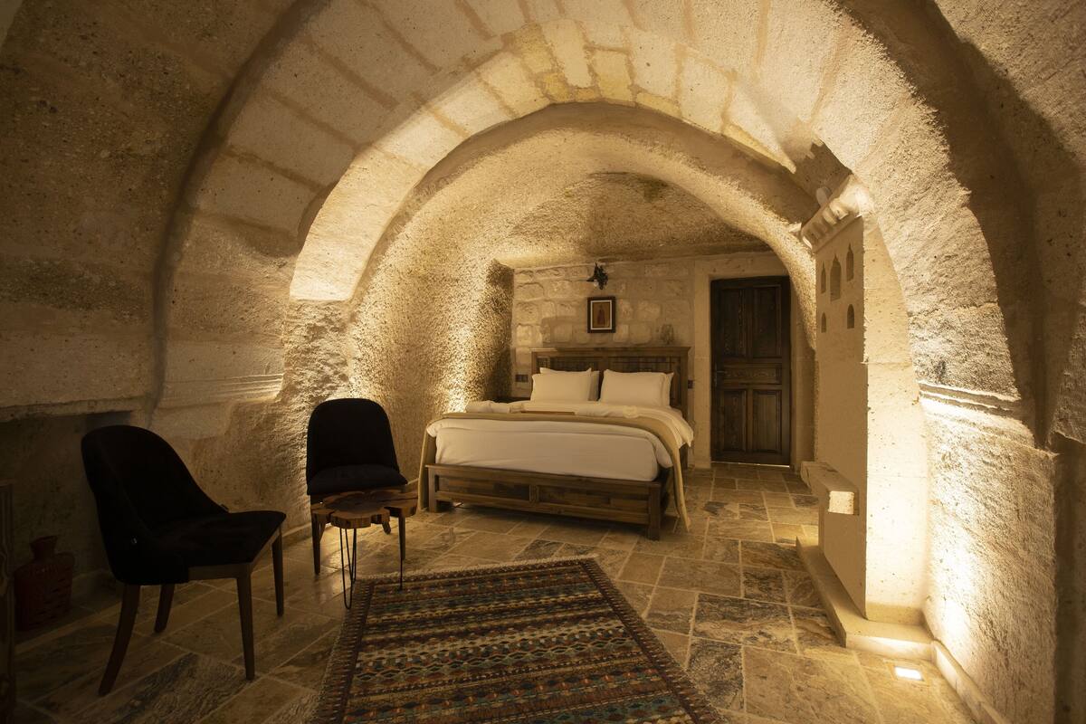A bedroom with a stone arched ceiling, a bed, two chairs, and a patterned rug.