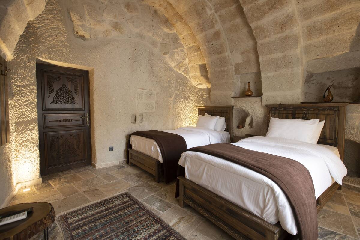 A stone-walled bedroom with two beds, a wooden door, and a nightstand.